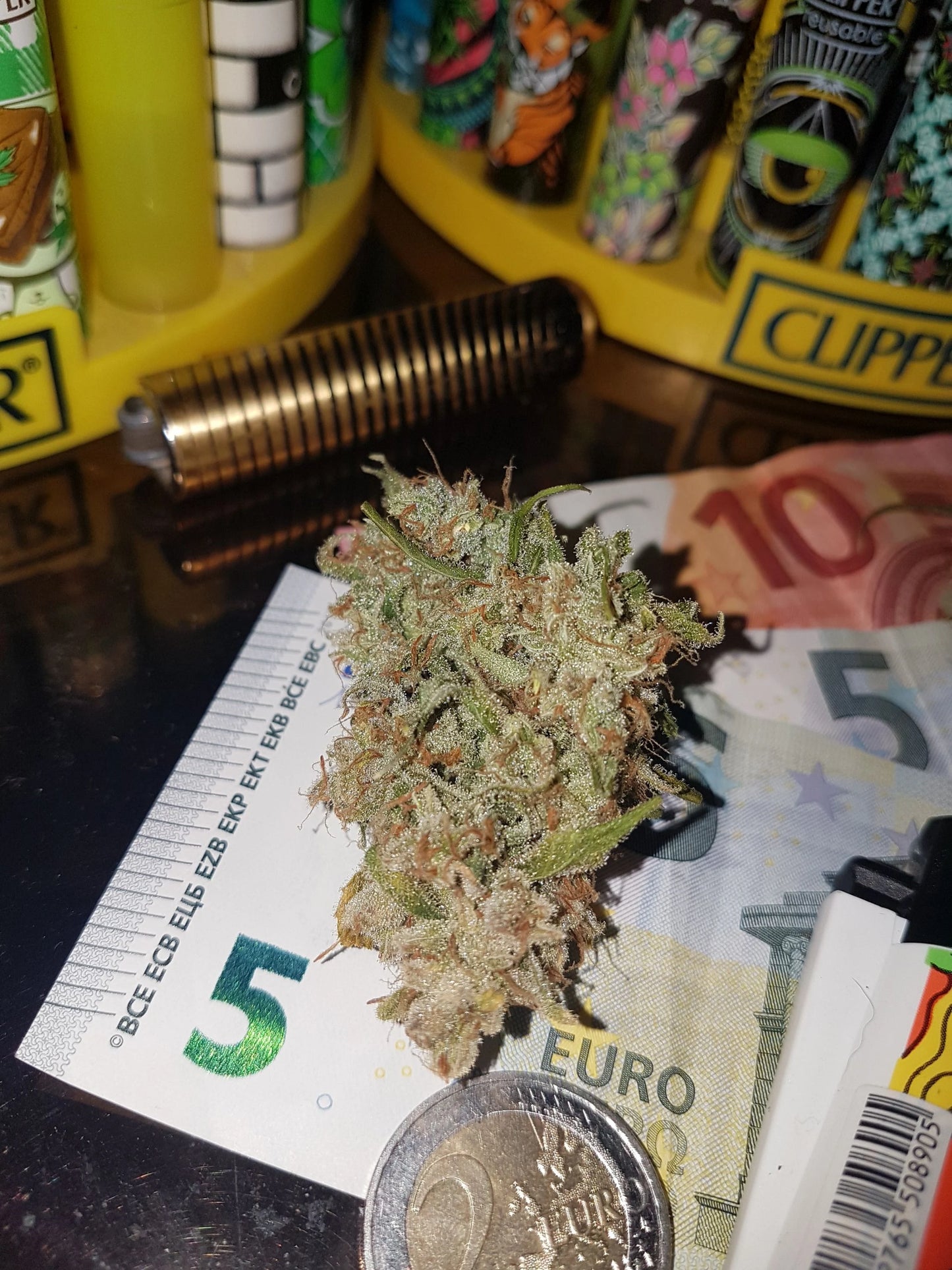 Close-up of a dense, resinous dried cannabis bud, covered in a glistening layer of trichomes. The bud is showcased against a Euro 5 note background, highlighting its intricate details and frosty appearance, typical of high-quality cannabis strains.