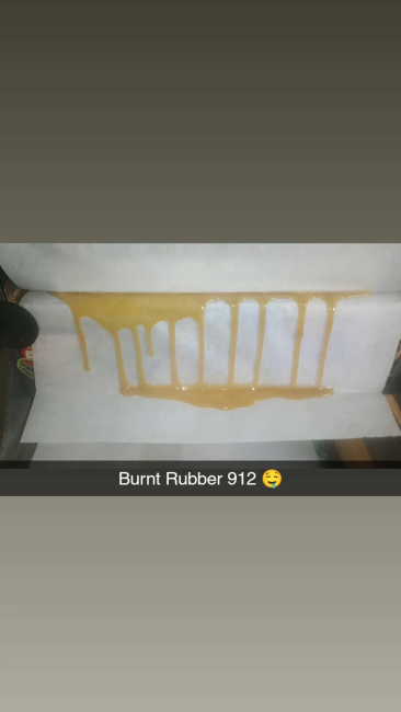 A square piece of white material, likely parchment paper or wax paper, featuring a horizontal smear of golden-brown burnt rubber. The image is framed with dark gray borders at the top and bottom, and a black bar at the bottom containing the text 'Burnt Rubber 912' with a laughing emoji.