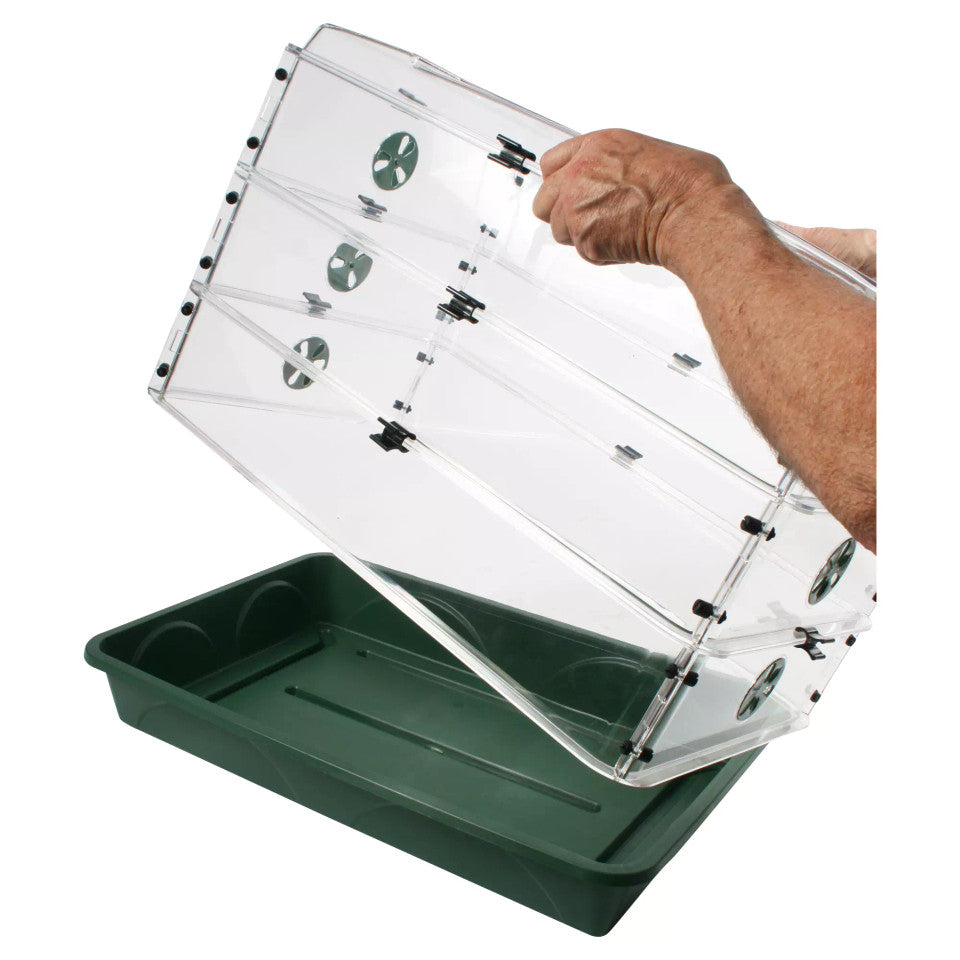 A clear acrylic raised garden bed cover with black clips being lifted by a person's arm over a dark green plastic tray, shown on a white background.